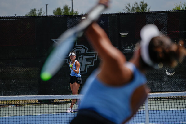 Two North Carolina women’s players practicing serves. One in the foreground out of focus has her left arm in the middle of a swing. Another in the background in focus is facing her teammate and getting ready to bounce a ball before her own serve.