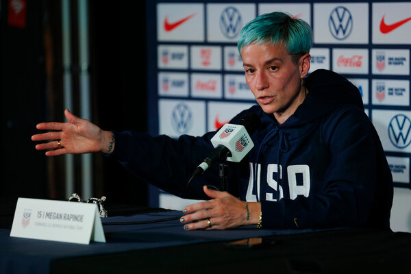 Rapinoe sitting at a table in front of a microphone, speaking and gesturing outward with her right hand.