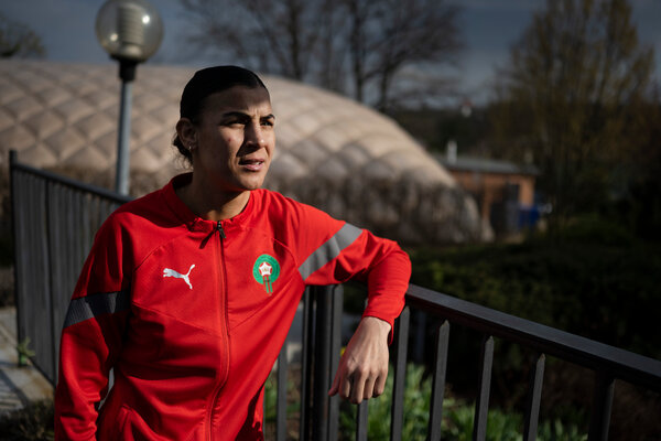 Khadija Rmichi, in a red warm-up jacket, leans up against a fence.