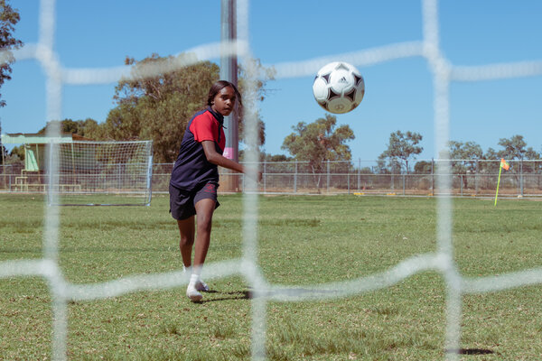 Shown from behind a soccer net, a girl watches her kick in midflight approaching the goal.