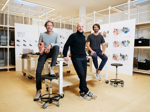 Three men pose for a portrait in a design studio with concept drawings of sneakers in the background.