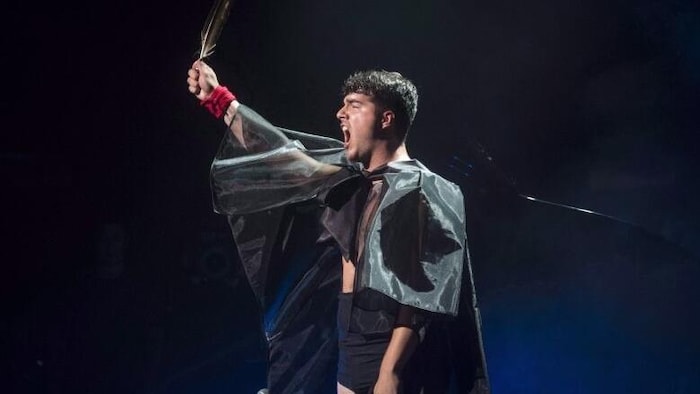 The singer sits in front of a microphone, raises his hand and shows a feather. 